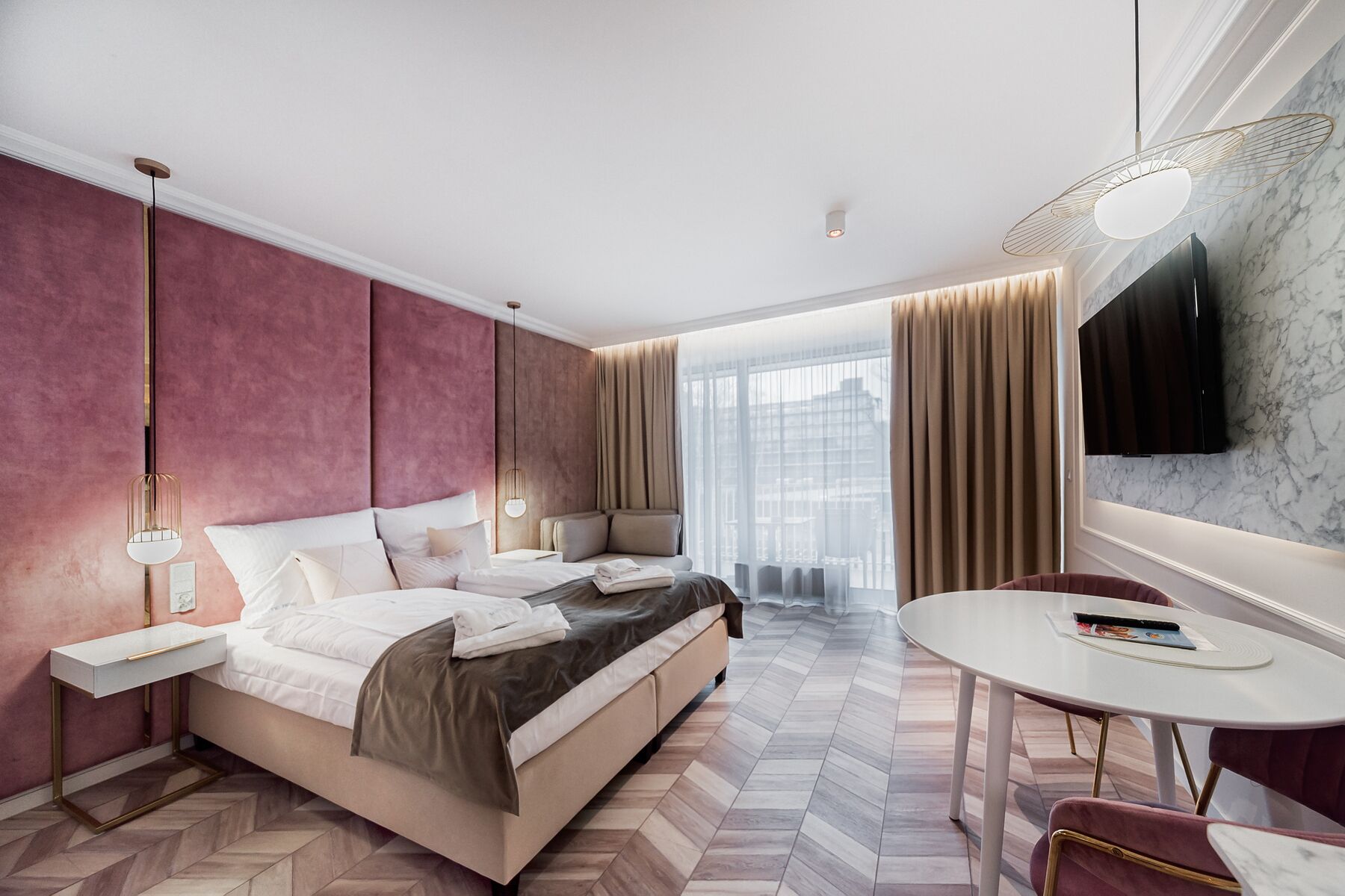 Una habitación de hotel moderna con una cama grande, un rincón de estar con una mesa y sillas, un televisor montado en la pared y vistas de la ciudad a través de grandes ventanales.