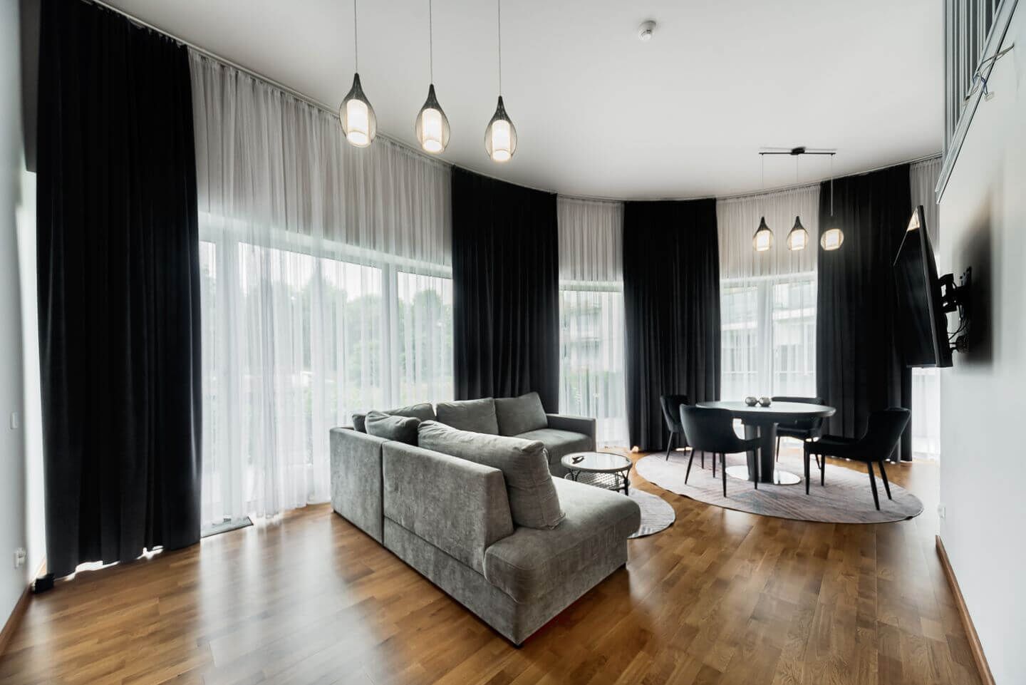 A modern living room with a grey sofa, a round dining table with chairs, and large windows with sheer curtains.