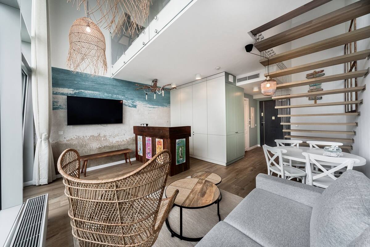 Ein modernes Wohnzimmer mit großem Fernseher, Rattanmöbeln, einer Treppe und Blick auf den Ozean.