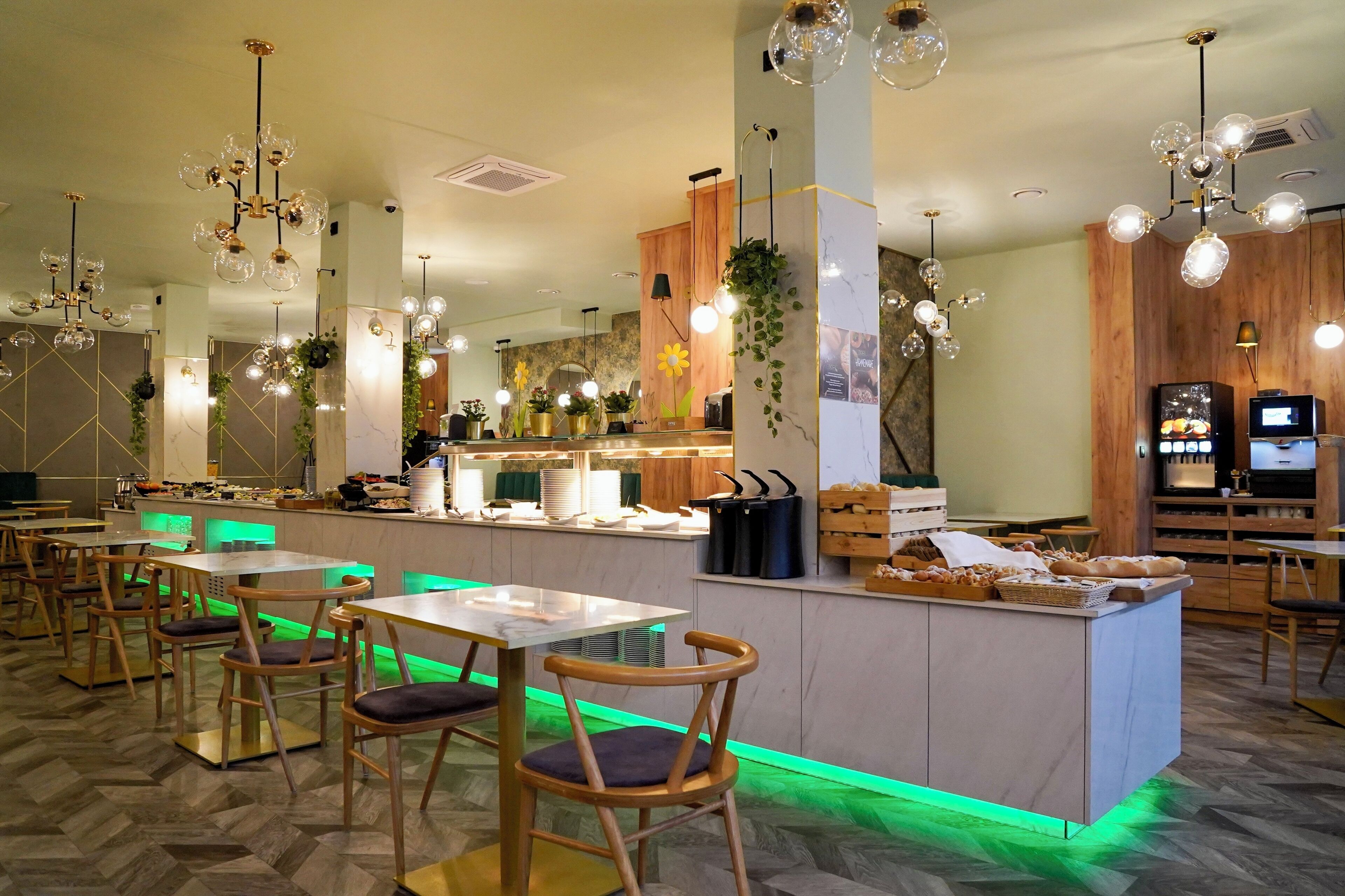 Um restaurante moderno com cadeiras de madeira, um balcão central com expositores de comida, luminárias esféricas pendentes e iluminação ambiente esverdeada.