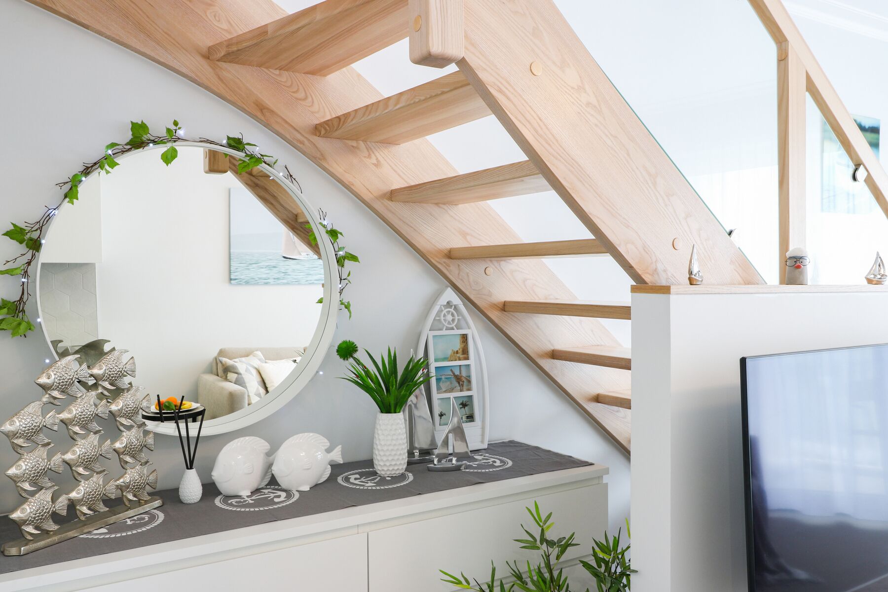 A modern interior with a wooden staircase, a round mirror with greenery, decorative fish, and a white console with a TV.