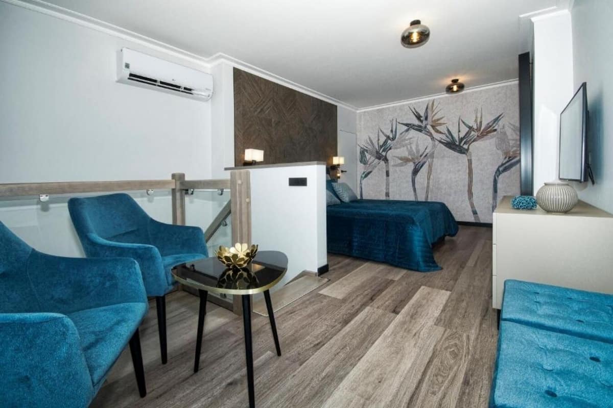 Une chambre d’hôtel moderne avec un plancher en bois, des fauteuils bleus, une petite table agrémentée d’un objet décoratif, un lit recouvert d’une courtepointe bleue, un climatiseur mural et une fresque représentant des bambous.