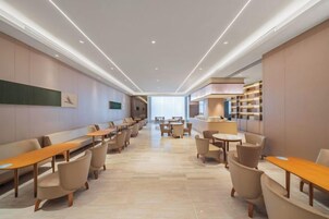 Un hall de hotel espacioso con mesas y sillas largas, una recepción y un amplio ventanal que proporciona luz natural.