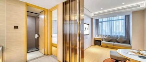 A modern hotel room with a wooden floor, a sofa set, a desk, and a large window with curtains.