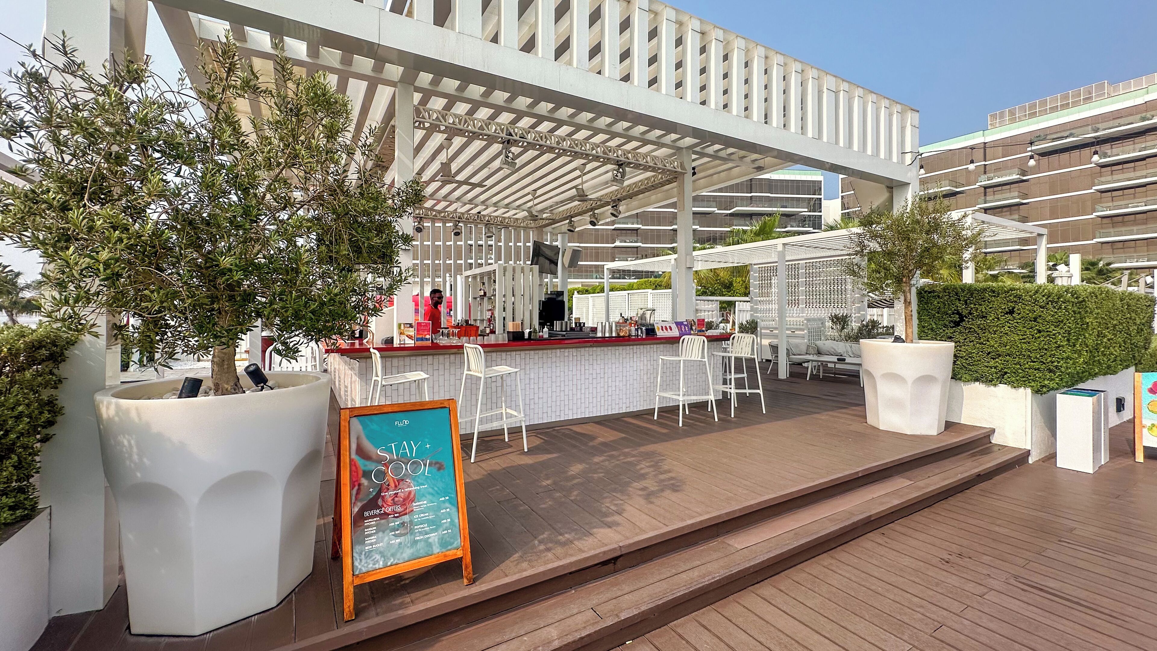 Un espace bar extérieur avec des tabourets de bar blancs, un panneau affichant le menu et un coin salon abrité.