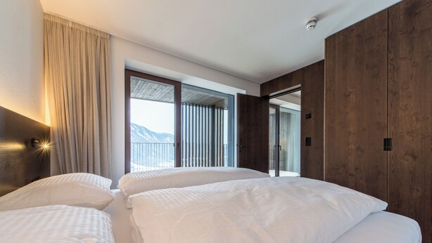 Une chambre d’hôtel avec deux lits, une grande fenêtre avec des rideaux, un lambris en bois et une vue sur les montagnes.