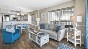 A spacious living room with a grey sofa, blue armchair, and a coffee table. A kitchen with white cabinets and a dining area with a blue table and chairs are visible. - Pelican's Perch @ Seawatch (Myrtle Beach)