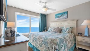 A bedroom with a large bed, a ceiling fan, a view of the ocean, a painting of seagulls, and a nightstand with a lamp. - Pelican's Perch @ Seawatch (Myrtle Beach)