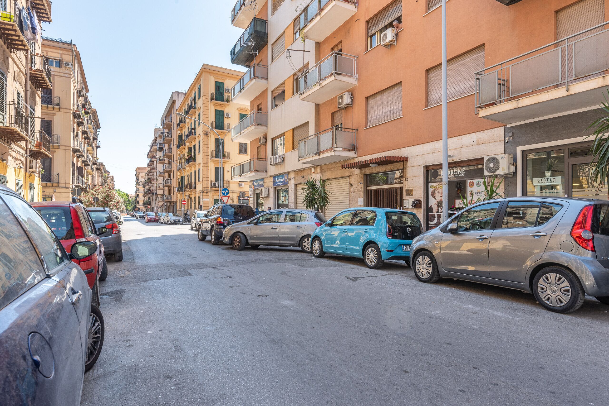 A street lined with multi-story residential buildings, parked cars, and a few storefronts.