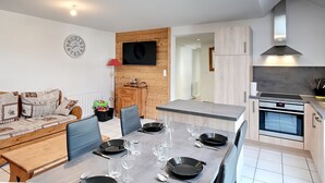 Une cuisine moderne dotée d’un four encastré, d’un coin repas avec une table grise et des chaises bleues, et d’un mur en bois sur lequel est fixée une télévision.