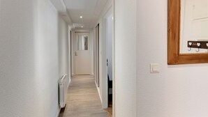 Un couloir étroit aux murs blancs, doté d’un plancher en bois et d’un radiateur blanc. Une porte se trouve au bout du couloir, et un tableau encadré en bois est accroché au mur.