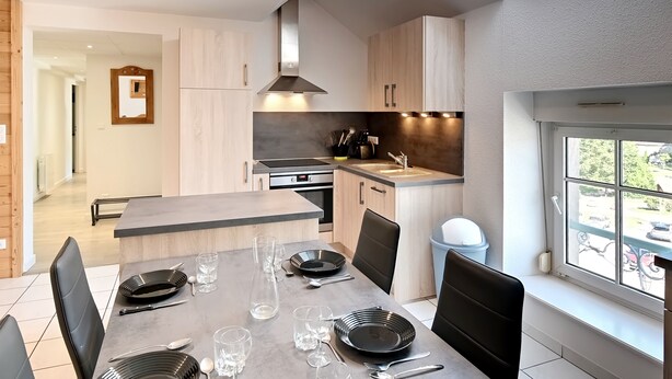 Une cuisine moderne dotée d’un îlot central, d’un coin repas avec une table dressée pour quatre personnes et d’une vue sur l’extérieur grâce à une grande fenêtre.