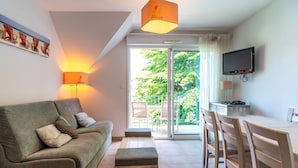 A living room with a sofa, ottoman, dining table, chairs, a TV, and a sliding glass door leading to an outdoor area. - Le Nid de Ker an Aod - Appt à 100m de la plage (Benodet)