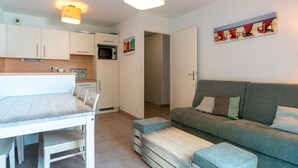 A modern kitchen with white cabinets, a dining table with chairs, a grey sofa, and a painting of beach chairs on the wall. - Le Nid de Ker an Aod - Appt à 100m de la plage (Benodet)
