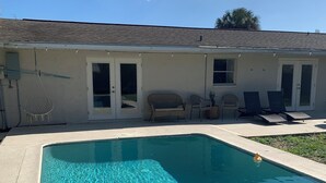 A rectangular swimming pool with clear blue water, a covered patio with outdoor furniture, and a building with a flat roof and sliding glass doors. - Pool Heated House - Near beach in Port Charlotte (Port Charlotte)