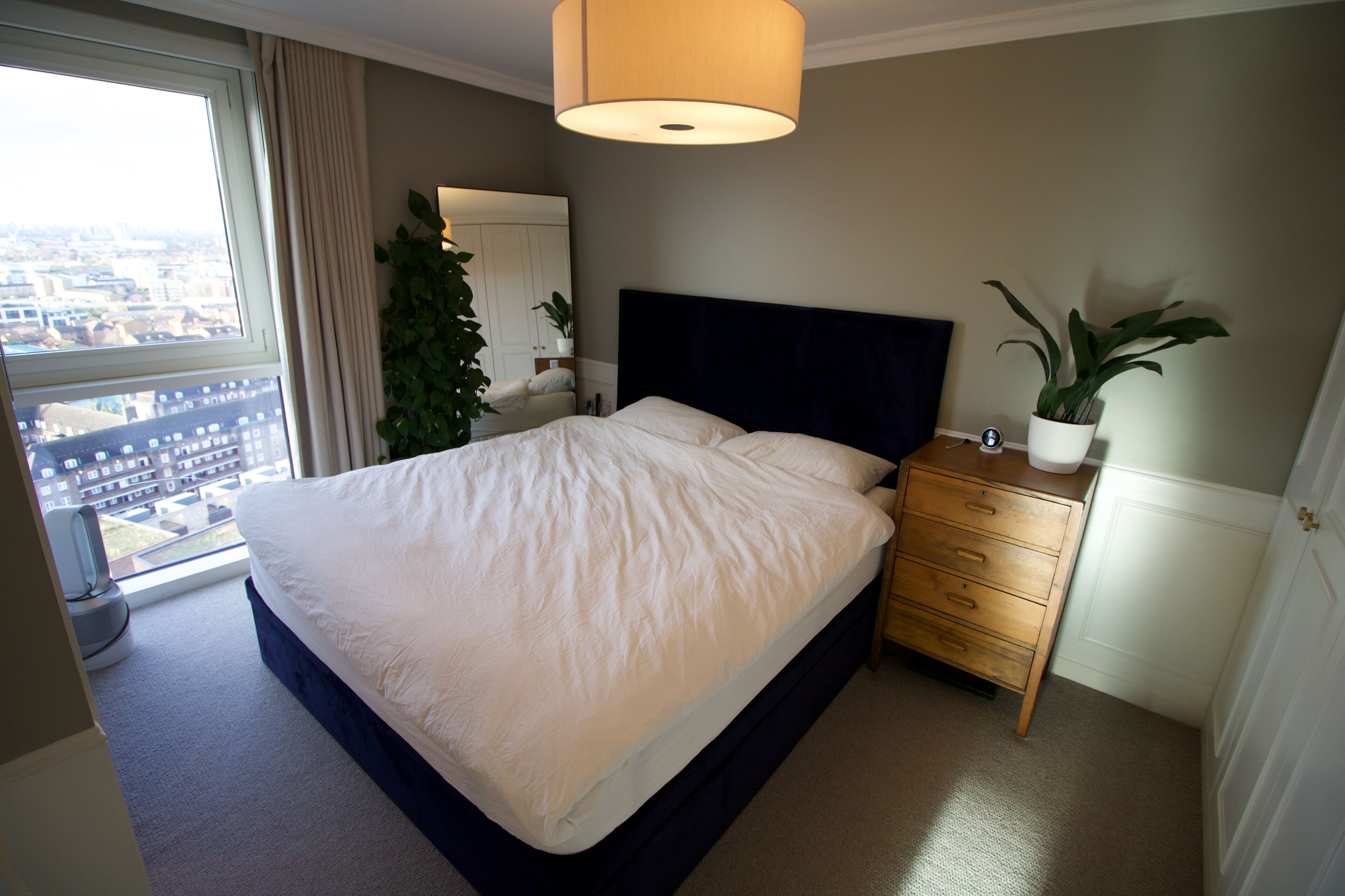 Une chambre avec un grand lit, une table de chevet, une commode, une plante en pot et une fenêtre donnant sur la ville.