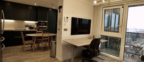 A modern kitchen with dark cabinets, a dining area with a round table and chairs, a large window with a city view, and a workspace with a desk and chair.