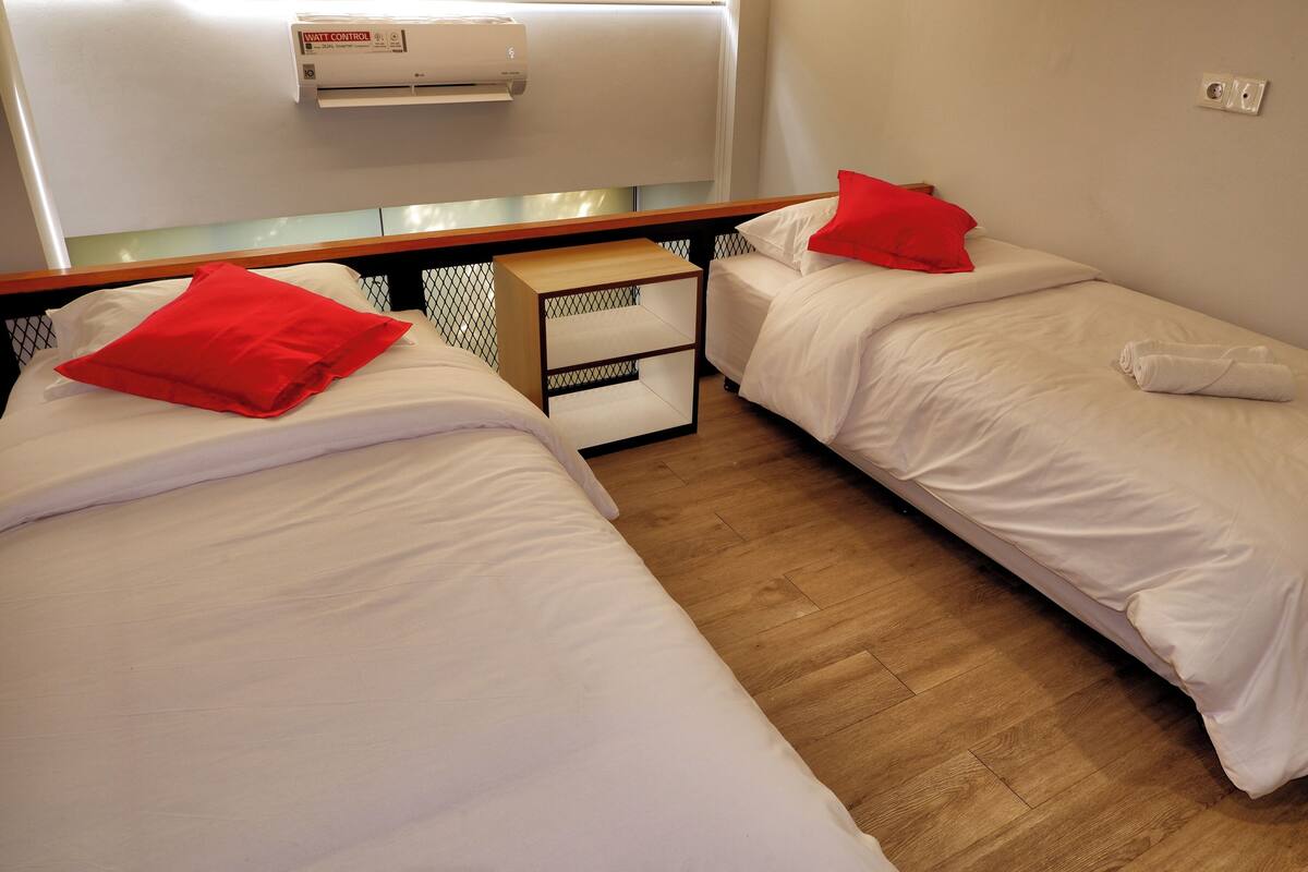 Ein Hotelzimmer mit zwei Betten, jeweils mit weißer Bettwäsche und roten Kissen. Zwischen den Betten steht ein Nachttisch aus Holz, und über den Betten ist eine weiße Klimaanlage an der Wand montiert.