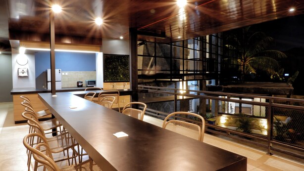 Un comedor moderno con una mesa larga y sillas de mimbre, una cocina al fondo y vistas a palmeras a través de amplias ventanas.