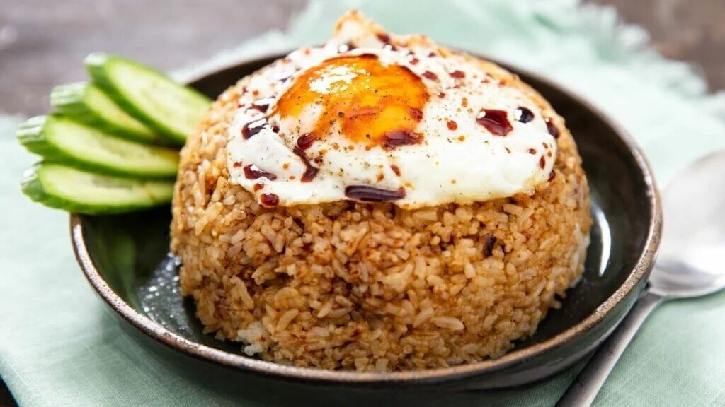 A bowl of fried rice topped with a fried egg, accompanied by sliced cucumbers.