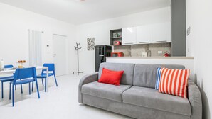 Un espacio habitable moderno con un sofá gris, almohadones azul y rojo a rayas, un área de comedor con sillas azules y una cocina con armarios blancos.