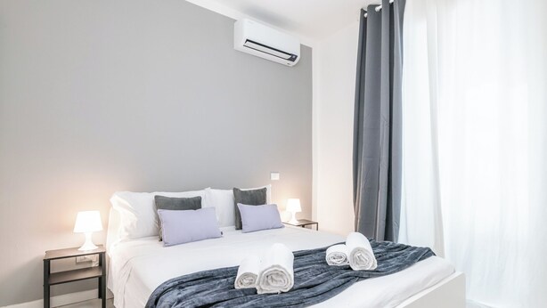 Una habitación de hotel moderna con una cama grande, dos mesitas de noche, aire acondicionado de pared y vistas al exterior a través de una ventana con cortinas.
