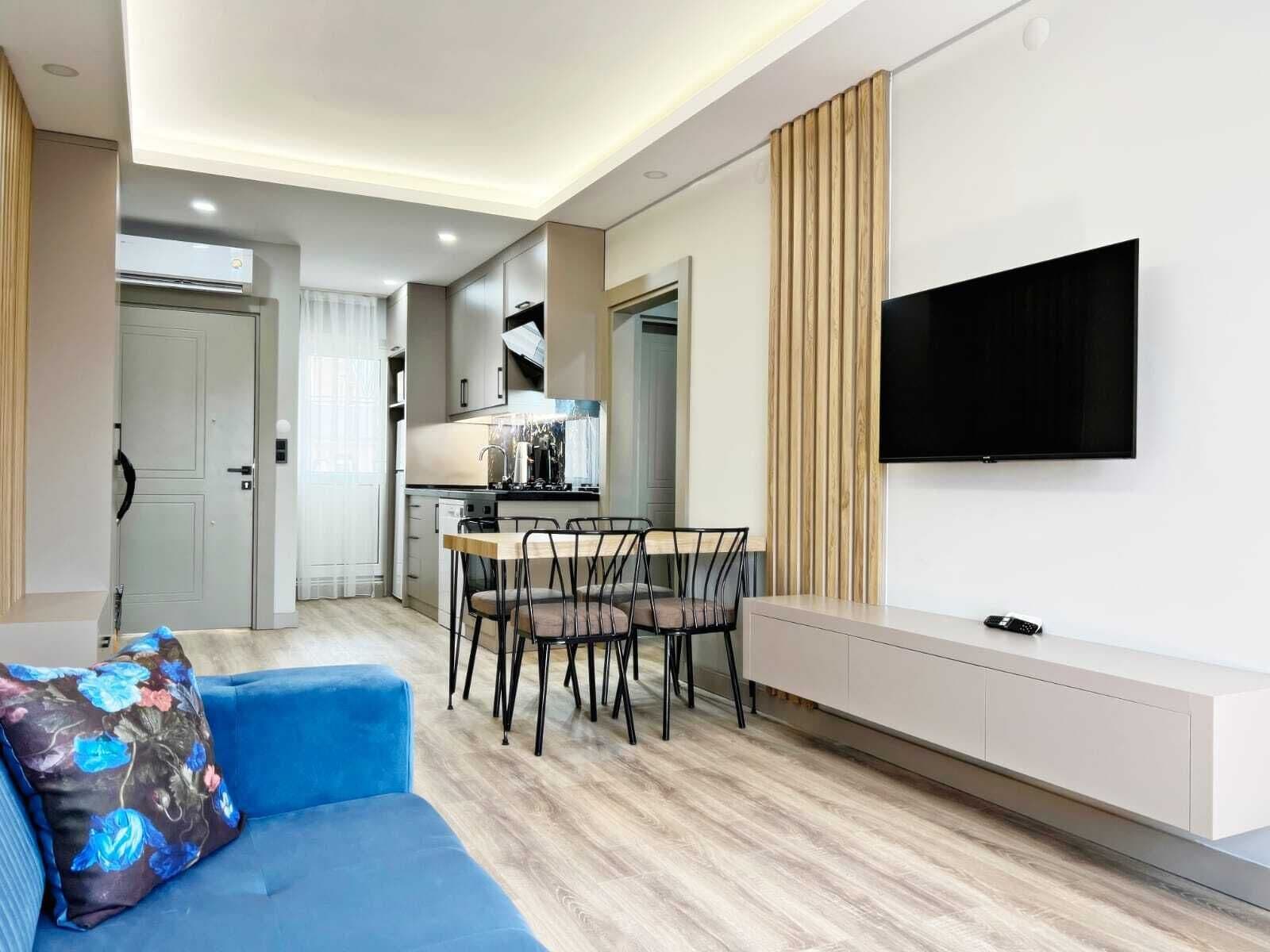 A modern living room with a blue sofa, a dining area with a wooden table and chairs, a kitchen in the background, and a TV mounted on the wall.