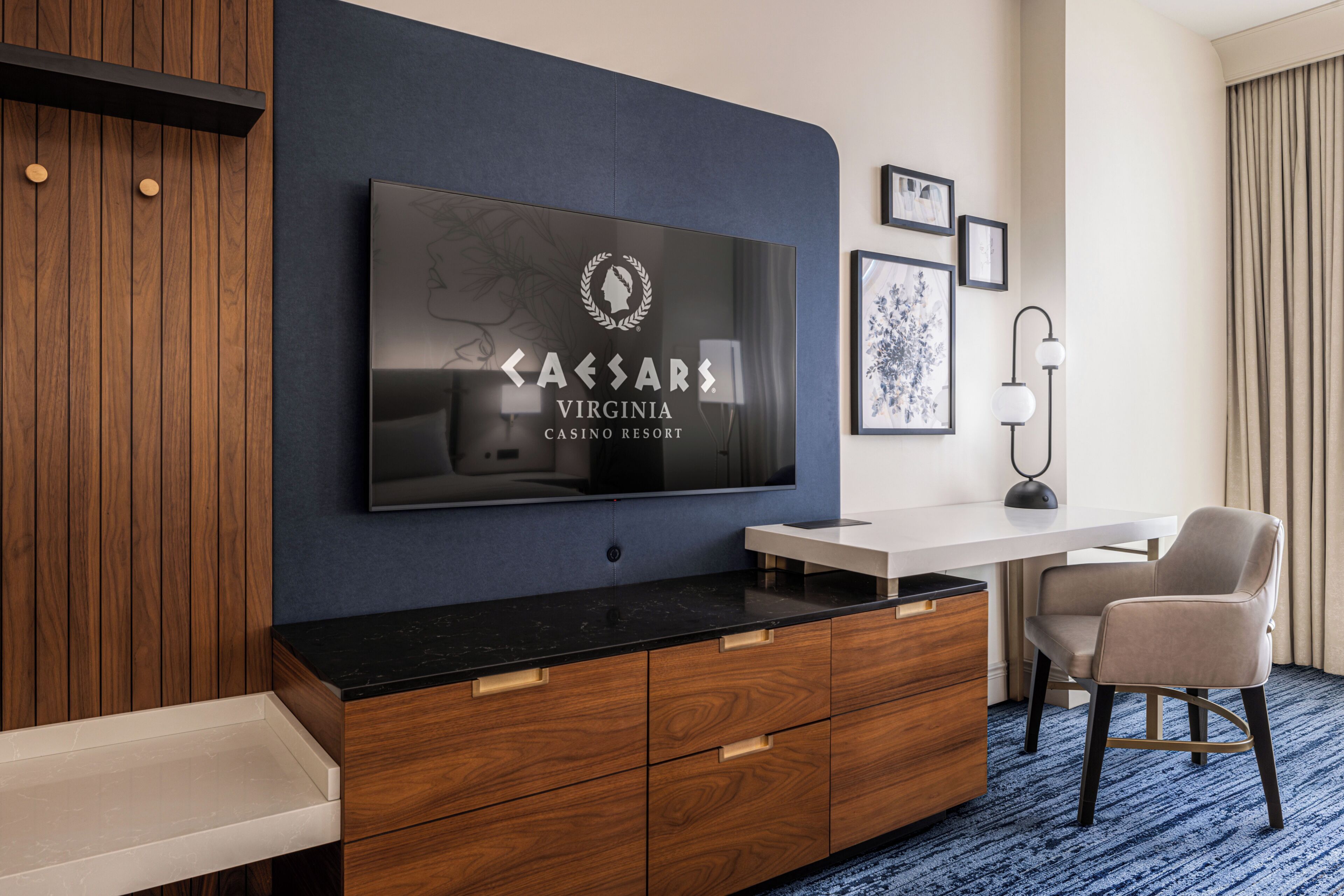Habitación de hotel moderna con televisor de pantalla plana montado en una pared azul oscuro, un mueble de madera con cajones, un escritorio blanco, una silla y una alfombra azul.