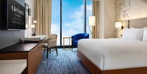 A hotel room with a large bed, a desk, a chair, a small table, and a view of the sky through large windows. - Caesars Virginia Hotel & Casino - A Caesars Rewards Destination (Danville)