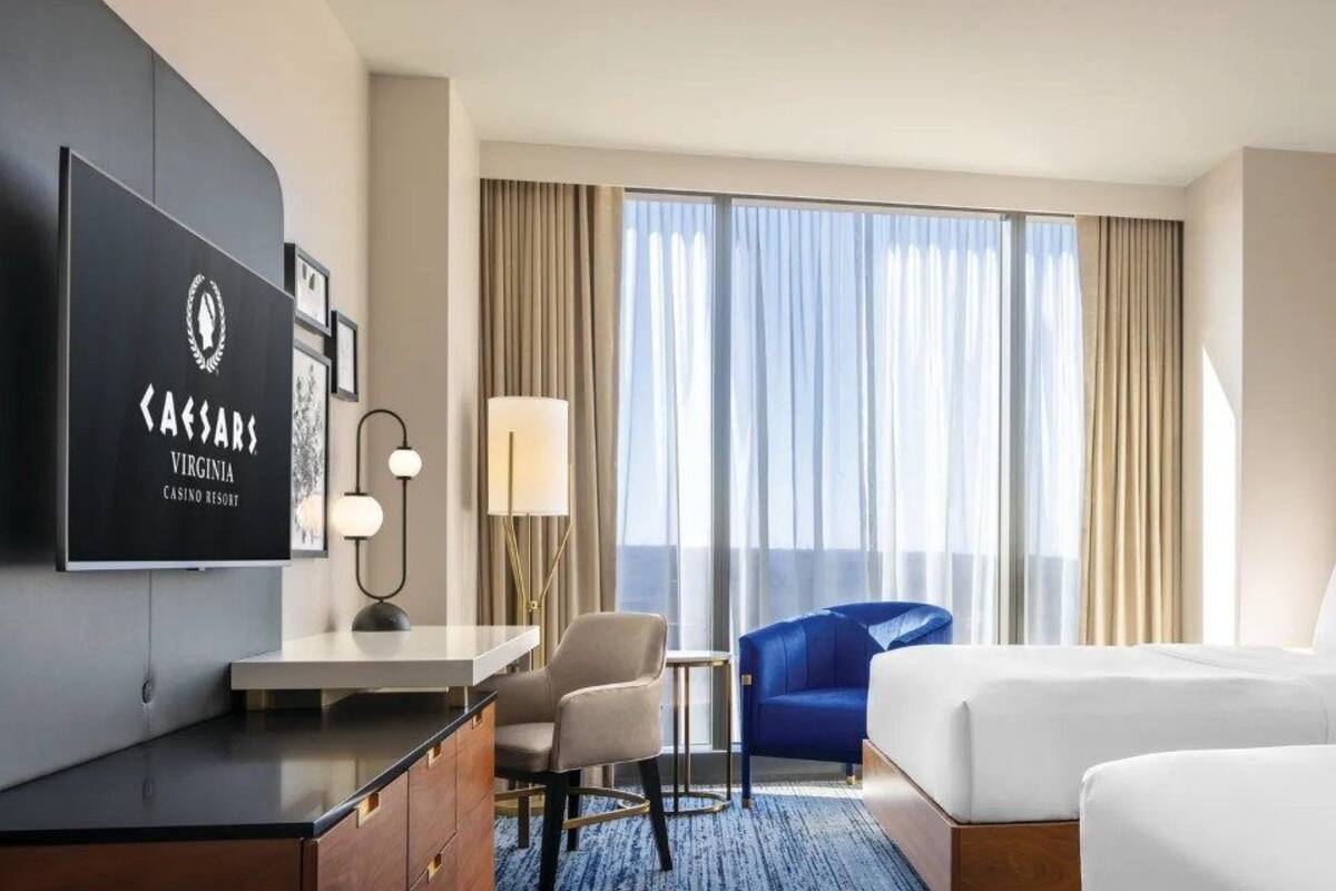 A hotel room with a TV, a desk, two beds, a chair, and a large window with curtains.