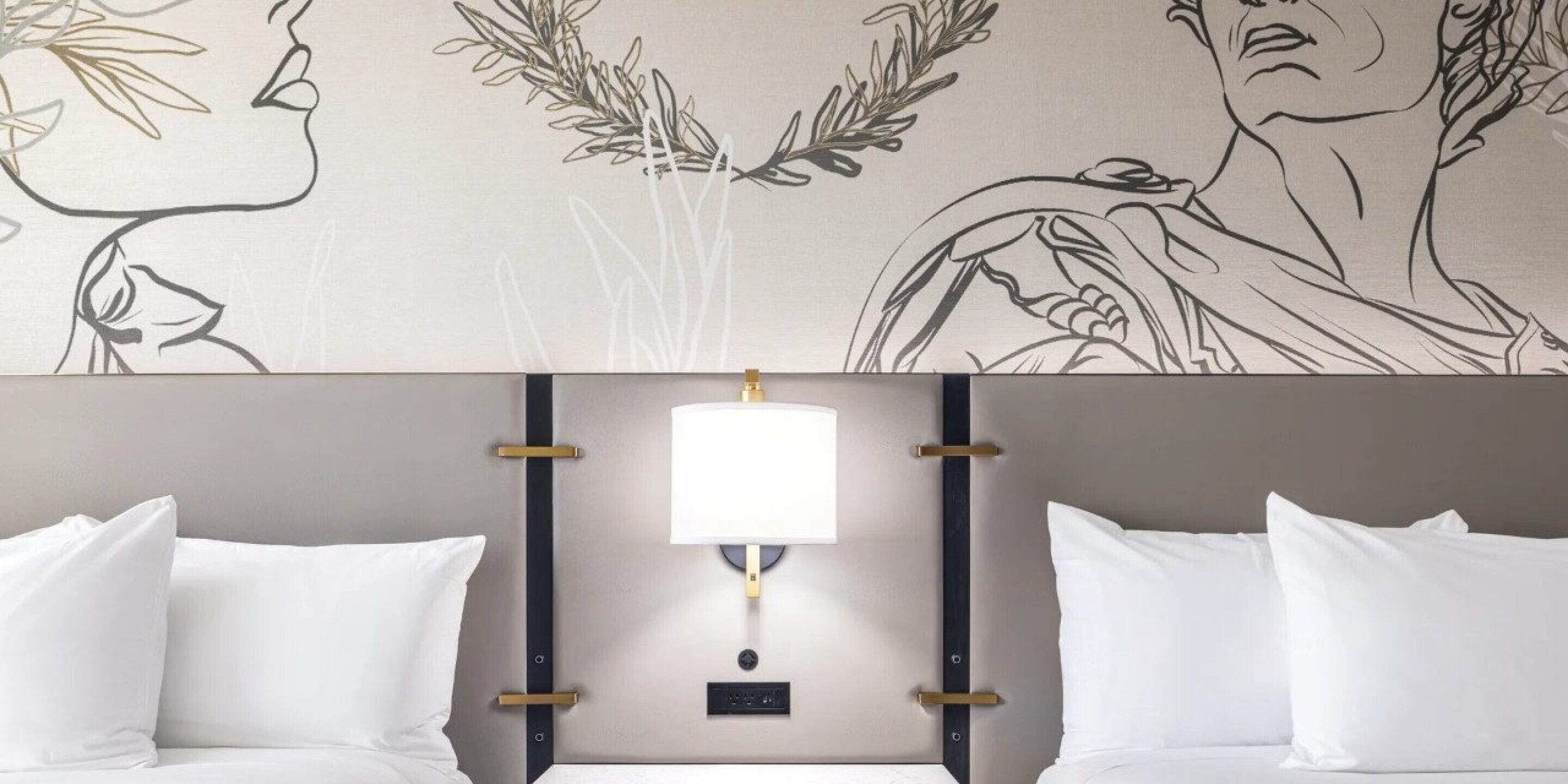 A hotel room with a bed, white pillows, a headboard with a light fixture, and a wall with a floral and portrait design.
