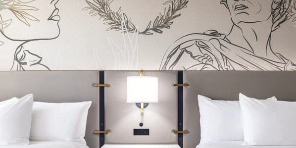 A hotel room with a bed, white pillows, a headboard with a light fixture, and a wall with a floral and portrait design.