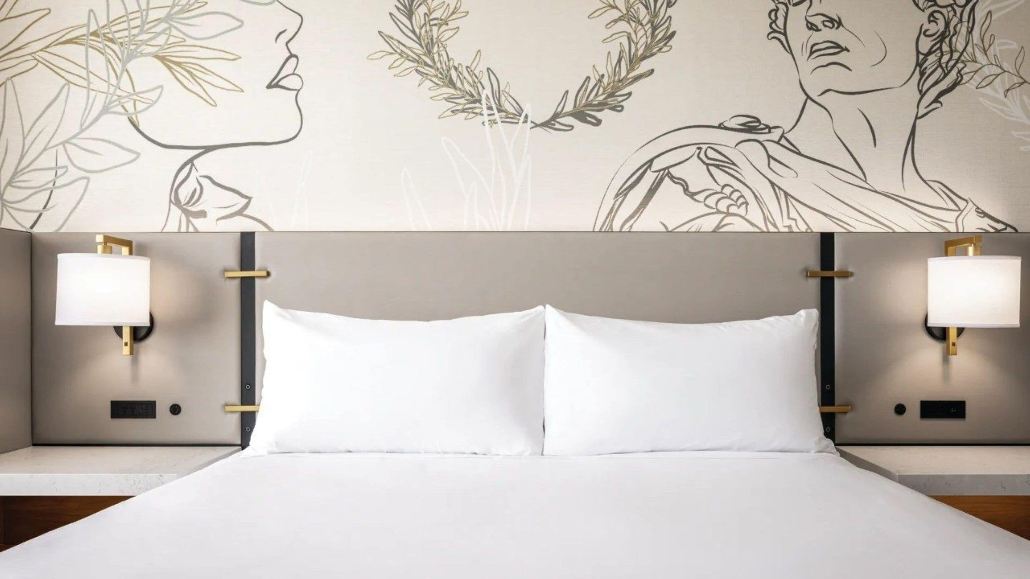 A bed with white linens, two bedside lamps, and a wall with a floral and portrait design.