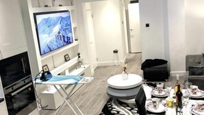 Een moderne woonkamer met een flatscreen televisie, een eethoek met een ronde tafel voor twee personen, en een keuken met een magnetron en oven.
