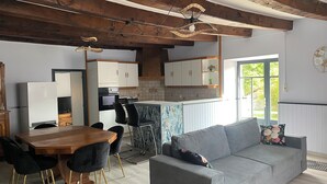 A modern kitchen with white cabinets, a dining area with a wooden table and black chairs, and a grey sofa in a living room with a view of greenery outside.