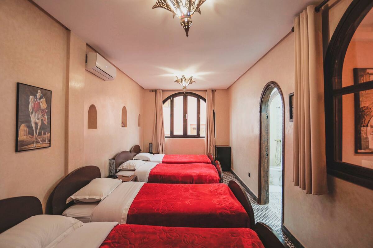 Quarto de hotel com duas camas, um candelabro, unidade de ar condicionado e vista através de uma janela arqueada.