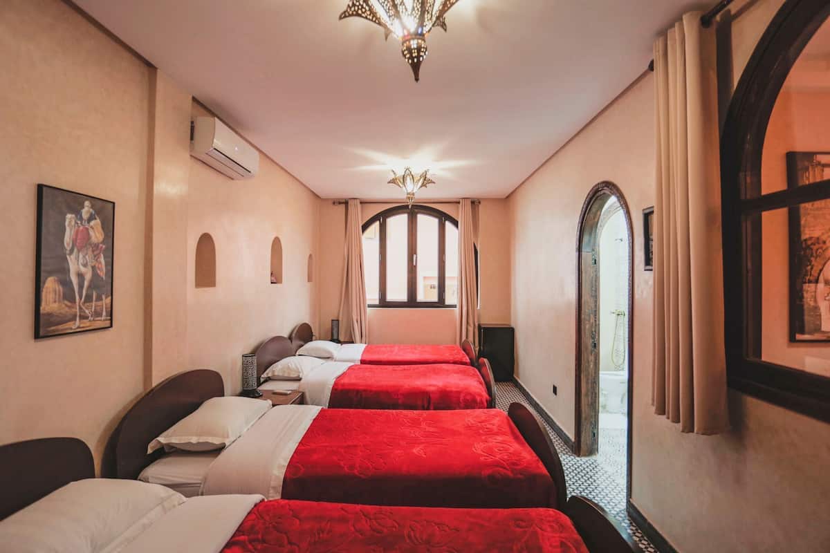 A hotel room with two beds, a chandelier, an air conditioning unit, and a view through an arched window.