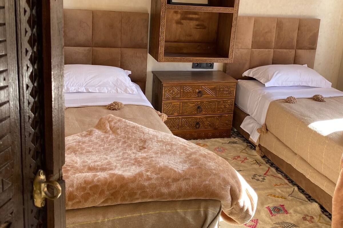 Quarto com duas camas, uma mesa de cabeceira de madeira com gavetas e uma prateleira sobre as camas.