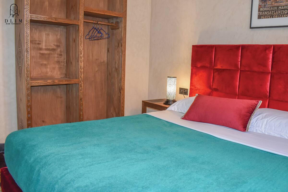 Cama com cabeceira vermelha, edredom azul-esverdeado e travesseiros brancos. Há um guarda-roupa de madeira com cabideiro e uma imagem emoldurada na parede.