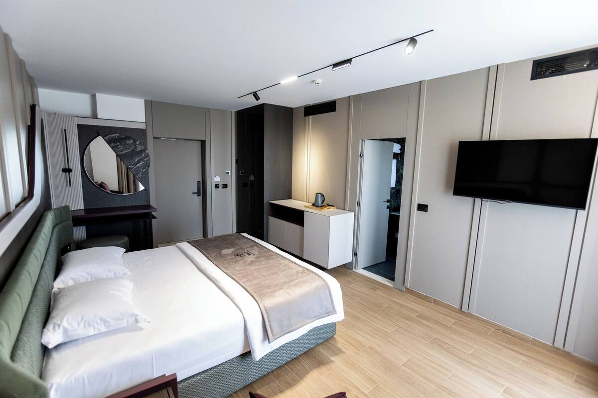 A modern hotel room with a large bed, a bedside table, a mirror, a TV mounted on the wall, and a bathroom visible through an open door.