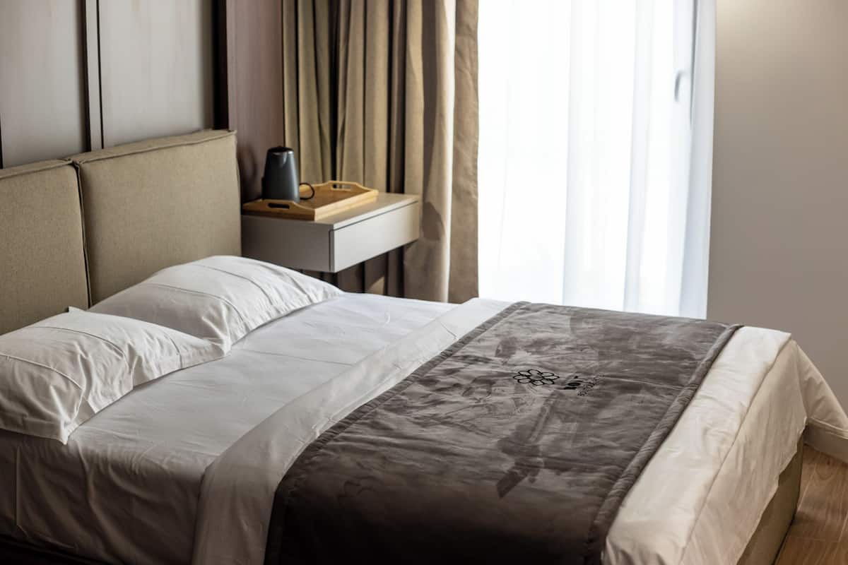 A neatly made bed with white linens and a gray throw blanket, a bedside table with a lamp and a tray, and a window with curtains.