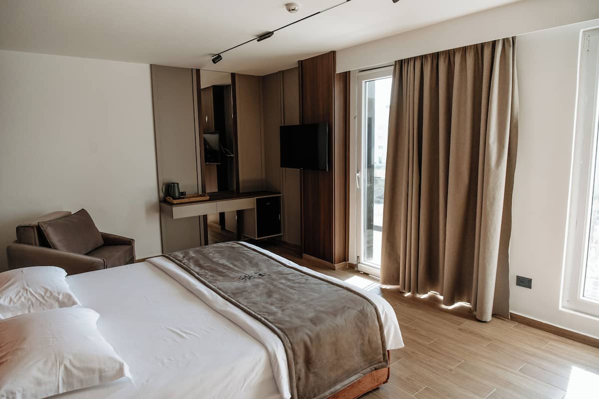 A modern hotel room with a large bed, a desk with a flat-screen TV, a chair, and a large window with curtains.