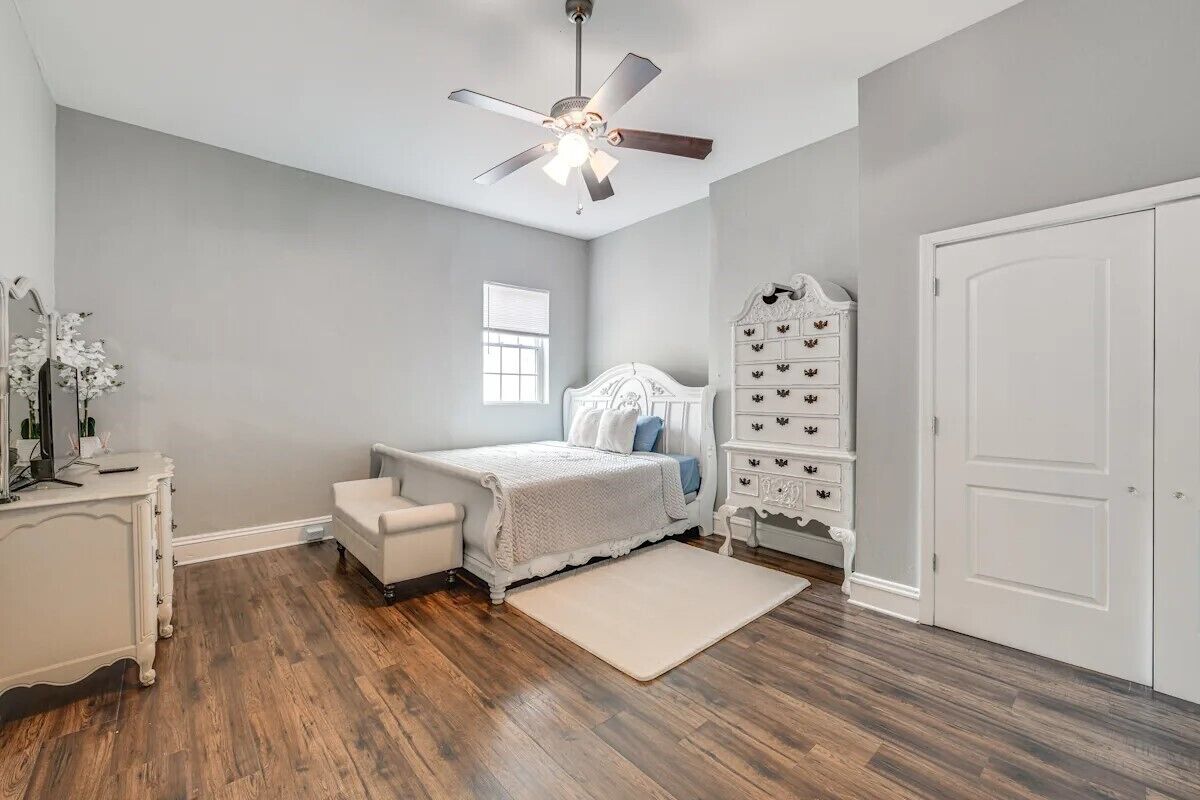 A bedroom with a bed, a dresser, a vanity, a ceiling fan, and a window.