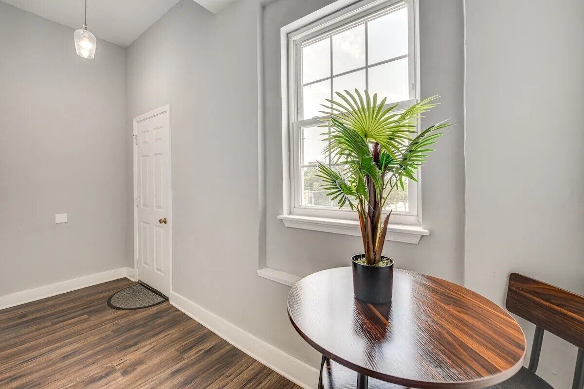Quarto com piso de madeira, uma mesa redonda com planta em vaso, uma janela com persianas e uma porta.