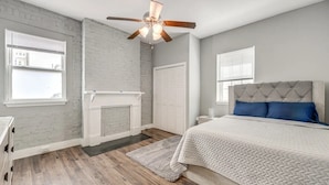 Un dormitorio con piso de madera, una cama blanca con almohadas azules, un ventilador de techo y una chimenea blanca.