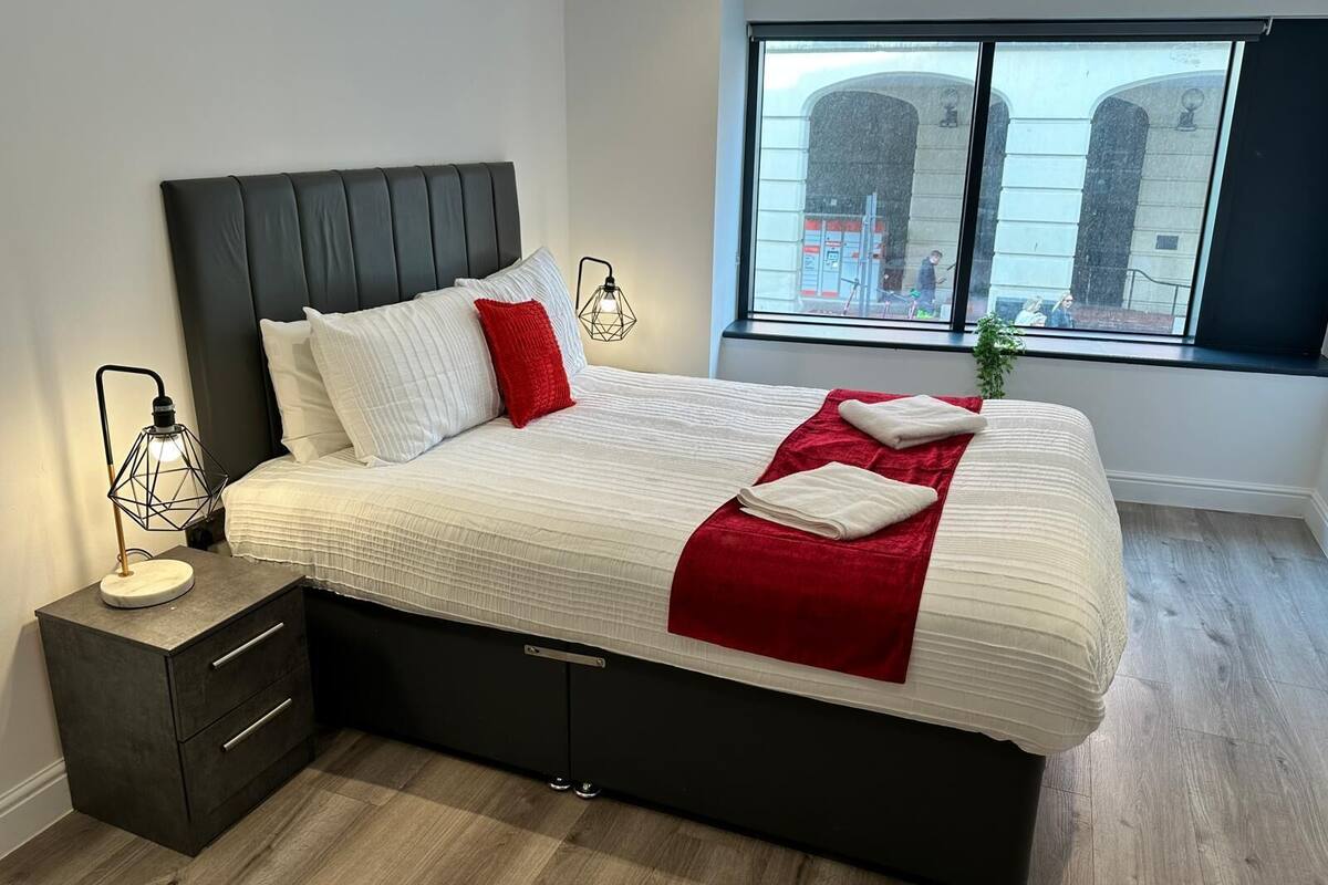 Une chambre moderne avec un grand lit, une table de chevet avec une lampe et une fenêtre donnant sur la ville.