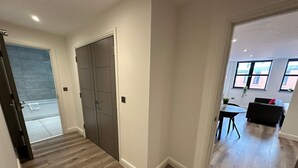 Un pasillo con suelo de madera, una puerta gris y vista a una sala de estar con un sofá negro y una mesa.