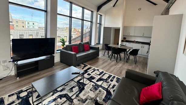 Una sala de estar moderna con un televisor grande, un sofá, una mesa de centro y un tapete estampado. Amplias ventanas ofrecen vistas a la ciudad.