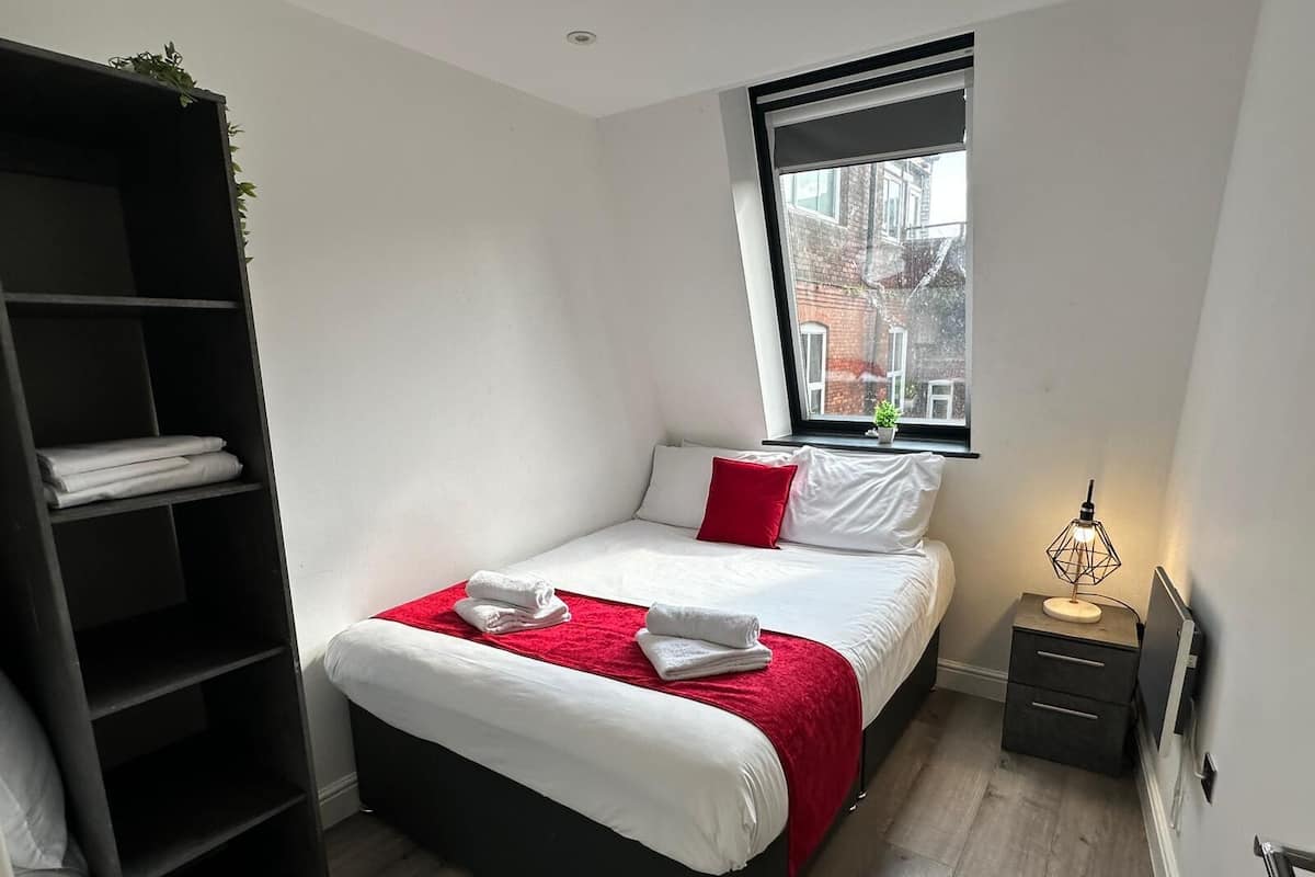 Une chambre à coucher avec un lit, un plaid rouge, une table de chevet, une lampe, une fenêtre et une étagère noire.