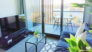 Ein modernes Wohnzimmer mit einem Flachbildfernseher, einem Glastisch, einer Couch und einem Balkon mit Blick ins Freie.